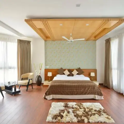 Spacious daylit master bedroom with a stepped maple-slat ceiling raft over the bed, a pale blue-and-gold damask wallpaper strip behind the wooden headboard