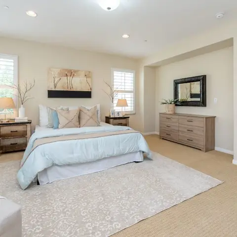 Spacious primary bedroom with a light-blue bedspread, matching wood nightstands with lamps, a wood dresser with mirror, and a patterned area rug over carpet.