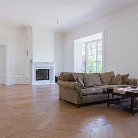 Sparsely furnished classical living room with white panel-moulded walls, a marble fireplace, chevron parquet flooring