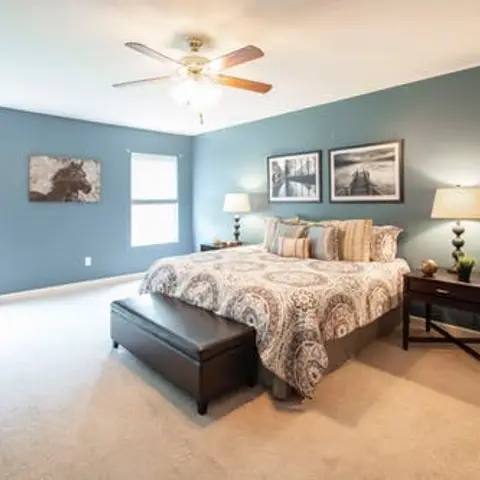 Staged bedroom with slate-blue walls, a patterned medallion bedspread flanked by matching table lamps, and a black leather bench at the foot of the bed.