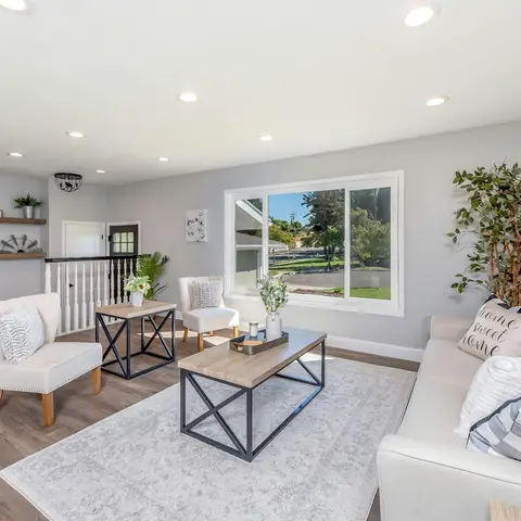 Staged living room with a beige tufted sofa and cream accent chairs around a black-metal-framed wood coffee table on a grey rug, a tall potted plant