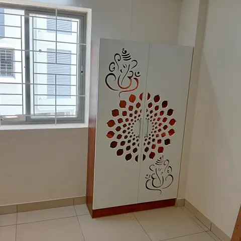 Standalone white pooja cabinet with laser-cut Ganesha motif and mandala jaali door panels on a wood-tone plinth base, set in an otherwise bare room.
