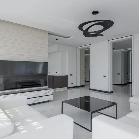 Stark white living room with an L-shaped white sofa, black glass coffee table and a horizontal stone-clad TV feature wall under a sculptural black ring pendant