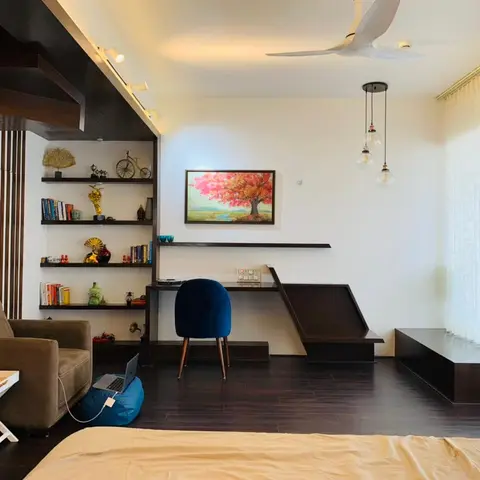 Study corner inside a bedroom, shot over the foot of the bed: wenge open shelving stacked with brass curios and paperbacks beside a cantilevered desk with a