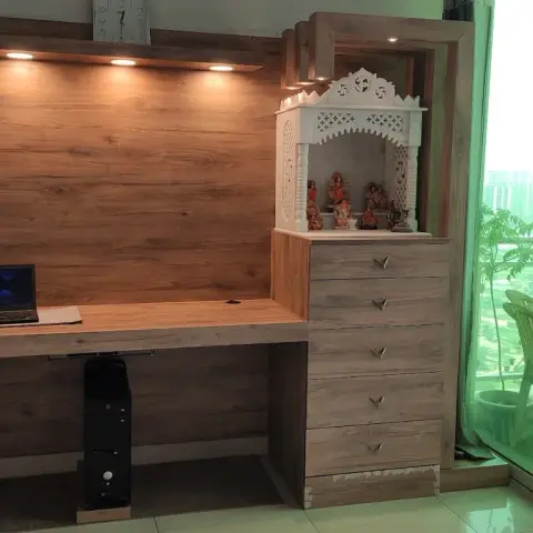 Study desk with a laptop setup beneath a wood-panelled overhead shelf, beside a white carved pooja unit built into a chest of drawers near the balcony.