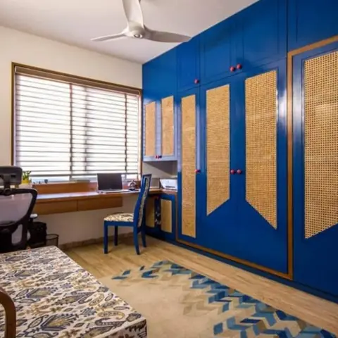 Study/home office with a floor-to-ceiling royal-blue wardrobe featuring rattan cane door panels and brass trim, a built-in wood desk with a mesh office chair