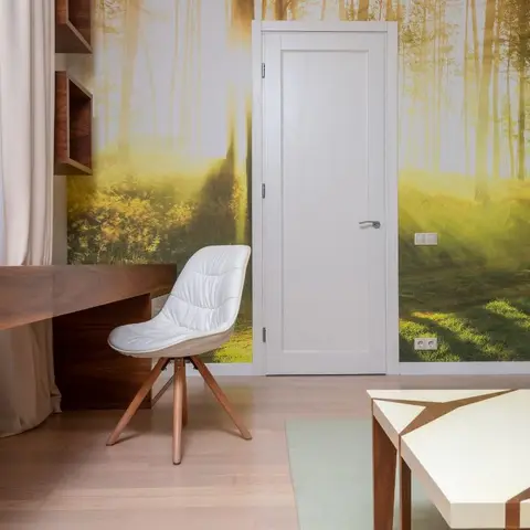 Study nook with a floating walnut desk and white cushioned chair against a floor-to-ceiling forest-mural wallpaper, beside a white door