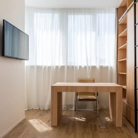 Study nook with a plain wood desk and chair by sheer-curtained windows next to a tall ladder-rail bookshelf.