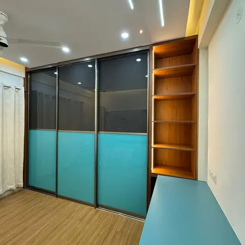 Study nook with a smoked-glass and teal sliding wardrobe beside a matching teal built-in desk with open wood shelving.