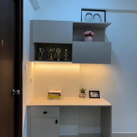 Study nook with a wall-mounted grey desk, backlit display shelving holding trophies and framed photos, and warm cove lighting beneath the cabinets.