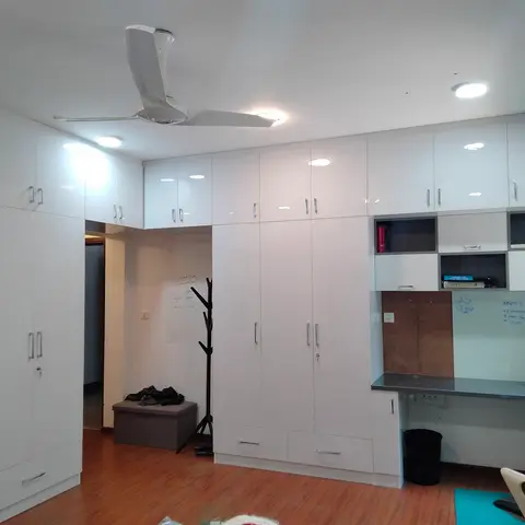 Study nook with a wall of glossy white wardrobes, a built-in desk with overhead storage niches, and a whiteboard for notes beside a coat stand.
