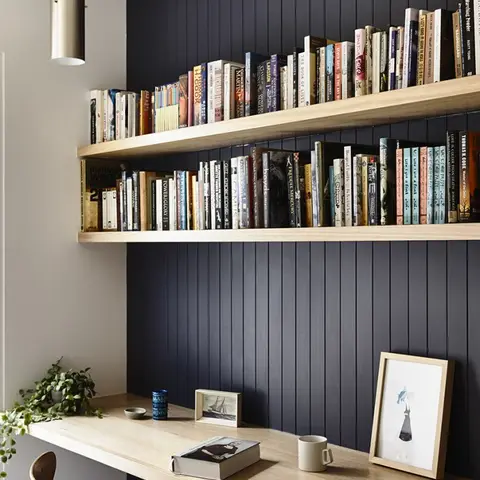 Study nook with navy vertical-slat wall panelling, two floating oak shelves stacked with books, a built-in oak desk and a cylindrical pendant light overhead.