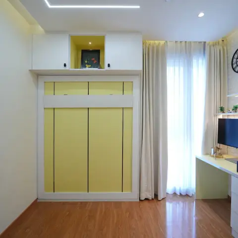 Study where a soft-yellow panelled floor-to-ceiling unit with a lit loft niche faces a white brick-clad wall of shelves above a corner desk with monitor