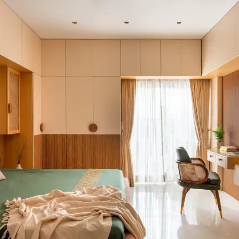 Sunlit bedroom where a beige wardrobe wall with a teak fluted dado wraps into a wall-hung study ledge, with a cane-and-leather chair