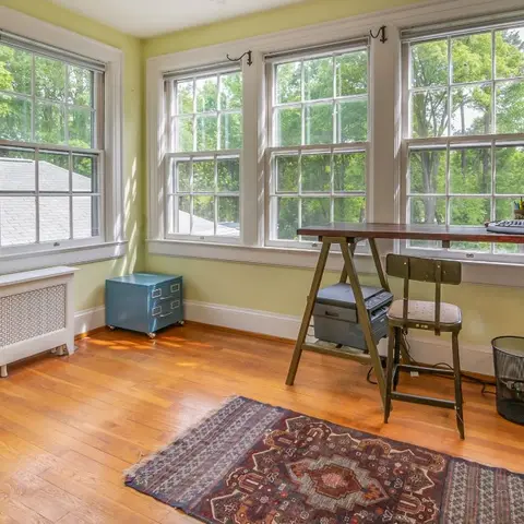 Sunlit home office with a trestle-leg desk and industrial task chair on a Persian-style rug, surrounded by multi-pane windows overlooking trees.