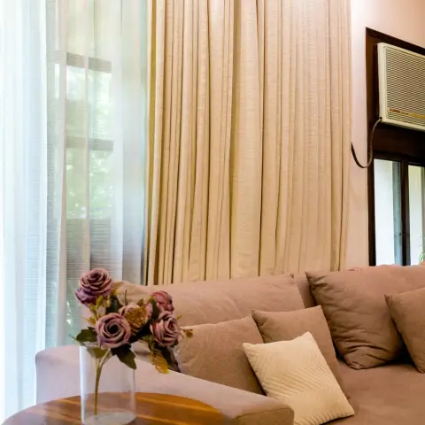 Sunlit living-room corner where floor-to-ceiling beige textured drapes layer over white sheers beside a taupe fabric sofa and a round teak-top side table