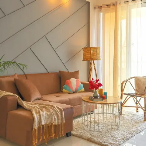 Sunlit living room with a rust-brown L-sofa against a grey wall of diagonal moulded panels, a cane tripod floor lamp and rattan chair by sheer curtains