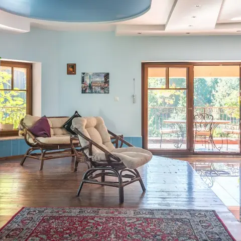Sunlit living room with pale blue walls, rattan lounge chairs on a patterned rug, and full-height glass doors opening onto a balcony.