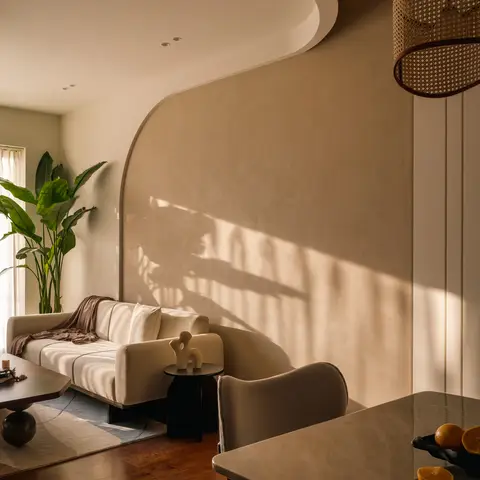 Sunlit open-plan space with a curved lime-plaster feature wall behind a beige sofa in the living zone and a marble dining table with a cane pendant light beyond