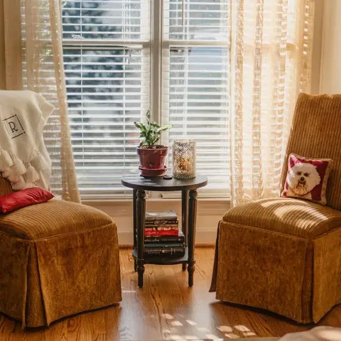 Sunlit reading nook with two skirted gold-chenille armchairs flanking a turned-leg side table stacked with books, in front of plantation-shuttered windows.