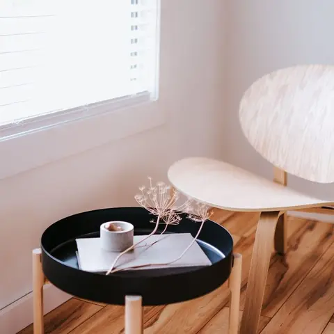 Sunlit room corner with a molded light-wood shell chair beside a round black-tray side table styled with a candle, notebook and dried flowers