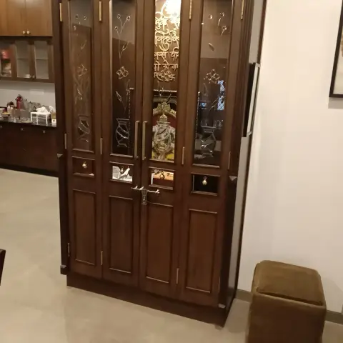 Tall teak mandir with etched-glass shutters carrying a gold shloka down the centre panel and a brass temple latch