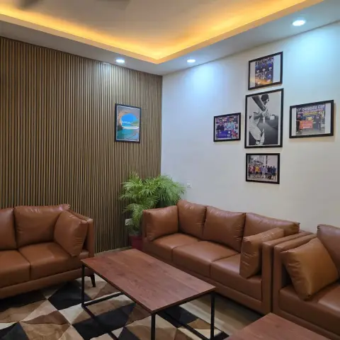 Tan leather 3+2 seater set against a full-height wooden slat wall, with a five-frame cricket photo gallery on the adjacent white wall and a black-and-tan