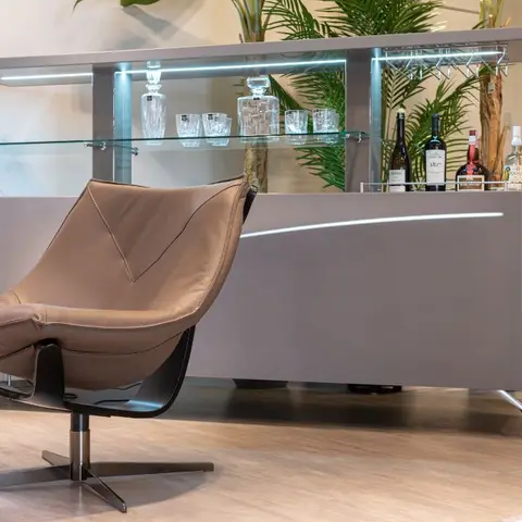 Tan leather swivel lounge chair beside a backlit home bar console with glass shelving, decanters and bottles