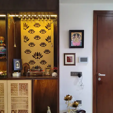Teak-veneer mandir with a lotus-cut gold back panel and a rail of brass bells, next to a framed Tanjore Lakshmi and a live-edge side table holding brass lotus