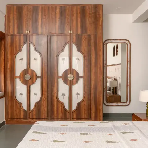 Teak wardrobe with mehrab-arched inset panels, palm-motif printed inserts and round brass pull plates, beside a rounded teak mirror