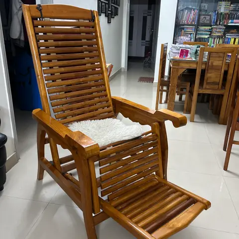Teak wood reclining planter's chair with a fold-out leg rest in an open living-dining area, with a glass-front curio cabinet and dining table visible behind.