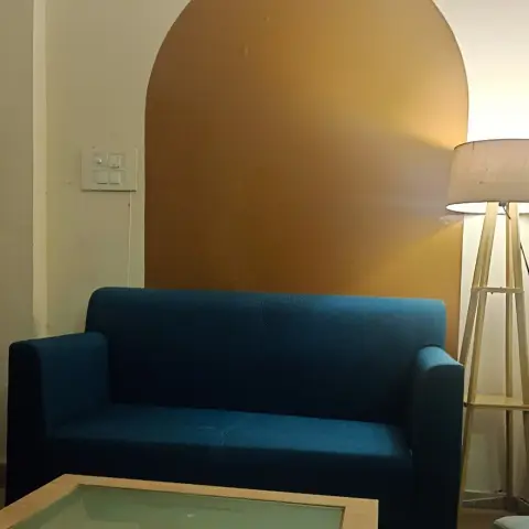 Teal two-seater sofa and a wood-and-glass coffee table set against a mustard arch-shaped colour-blocked accent wall, lit by a tripod floor lamp.