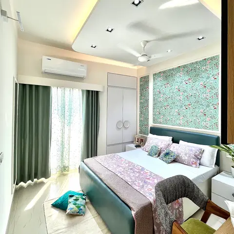 Teal-upholstered bed set against a teal floral wallpaper panel, with a white wardrobe, patterned curtains, a jute rug and a green velvet accent chair.
