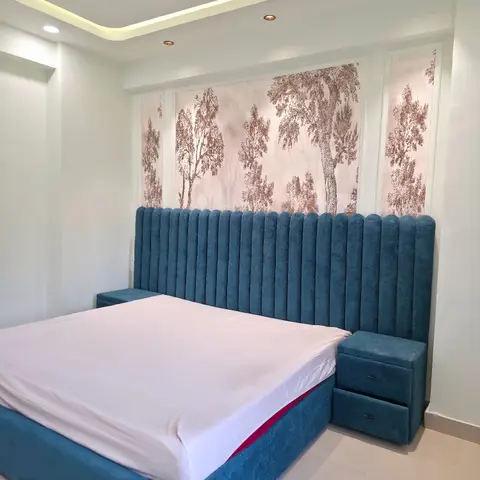Teal velvet channel-tufted headboard running wall to wall behind a low bed, with matching upholstered two-drawer bedside boxes and a sepia tree-print wallpaper