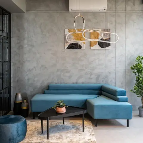 Teal velvet sectional set against a wall of grey concrete-finish slabs, with a looping brass-and-glass tube pendant hung across a two-panel ochre-and-black