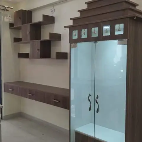 Temple-shaped wooden pooja cabinet with glass doors and brass hardware beside an asymmetric floating shelf unit, under a cove-lit ceiling.
