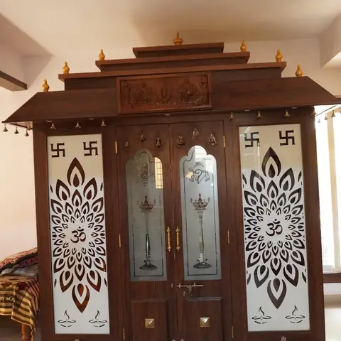 Temple-style wooden pooja mandir with a tiered dome roof, etched OM-motif glass door panels, and hanging brass bells.