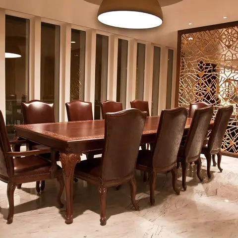 Ten-seat carved wood dining table with burgundy leather chairs on a marble floor, beside an ornate gold jaali partition screen.