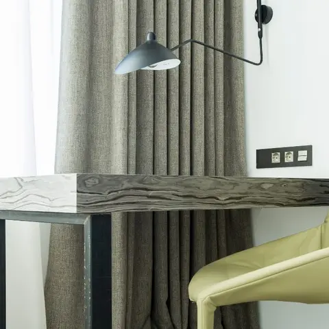 Tight corner view of a waterfall marble-and-wood console desk with a black articulated wall lamp and a pale leather armchair against grey curtains.