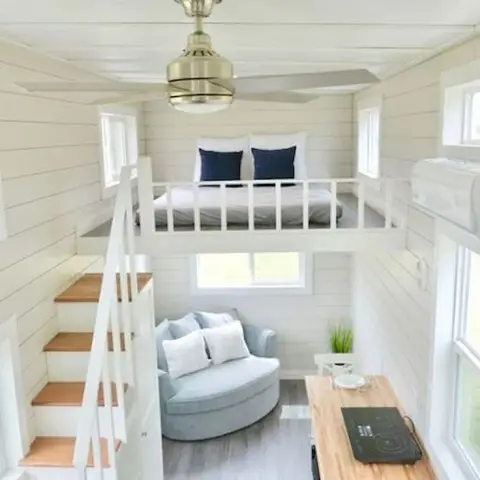 Tiny-house interior where a sleeping loft behind a white spindle rail sits over a round pale-blue loveseat