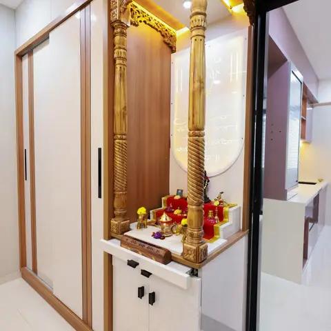 Traditional carved-wood pooja unit with backlit turned pillars and a red altar cloth, set beside a sliding-door wardrobe in the entryway.