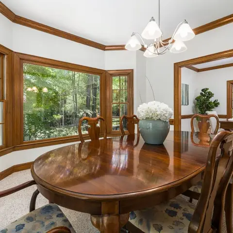 Traditional dining room with a bay window onto trees, stained wood crown moulding, and an oval mahogany table set with floral-pattern chair seats.