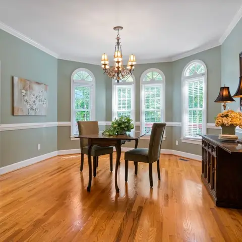 Traditional dining room with sage-green walls, arched windows, a wrought-iron chandelier, and a wood pedestal table with upholstered chairs.