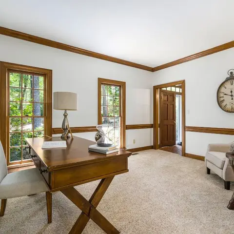 Traditional home study with a wood X-leg writing desk and upholstered chair by a window, a decorative oversized wall clock