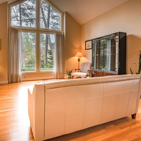 Traditional living room with a vaulted ceiling and tall arched window onto trees, a cream leather sofa facing a dark wood glass-front display cabinet.