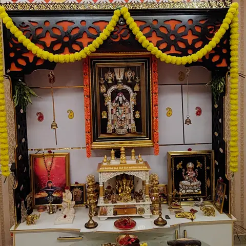 Traditional pooja unit with an orange-and-black carved jaali canopy and marigold garlands framing a marble-finish mandir with brass deity idols