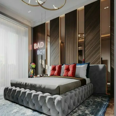 Tufted grey velvet bed against a vertical dark-wood panelled wall with brass trim, red and navy cushions, and a neon 'BAD BOY' sign beside the nightstand