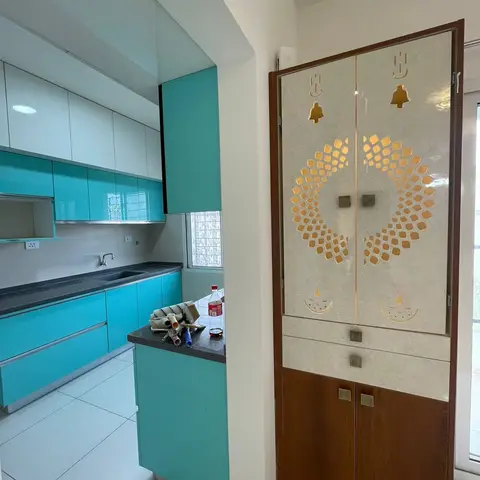 Turquoise glossy modular kitchen viewed past an ornate cream-and-wood pooja cabinet with laser-cut om, bell and diya motifs on its doors.