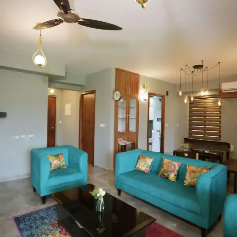 Turquoise sofa and armchairs with floral cushions on a distressed magenta-and-blue rug, brass teardrop pendants hanging beside a copper-hub ceiling fan