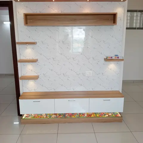 TV feature wall with book-matched white marble cladding, floating wood display shelves, and a backlit pebble-filled base beneath white drawer units.